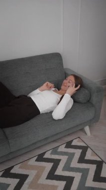 Genç bir kadın yeşil kanepede rahat bir şekilde uzanıyor, telefonla konuşuyor, sesli mesaj dinliyor ve akıllı telefonunda geziniyor, rahatlamış ve oturma odasında dikey görüntüsü var.