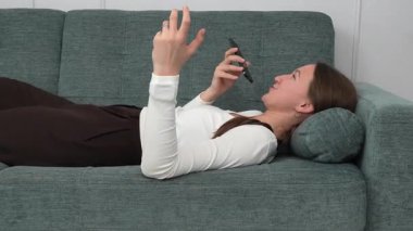 Beyaz gömlekli ve kahverengi pantolonlu genç bir kadın rahat bir koltukta uzanıyor, akıllı telefonu sağ elinde tutuyor ve bir sesli mesaj kaydediyor.