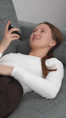 Gri kanepede dinlenen genç bir kadın, akıllı telefonla arkadaşlarıyla mesajlaşırken aynı zamanda kahkahadan konsantrasyona kadar olan dinamik bir duygu yelpazesini gösteriyor.