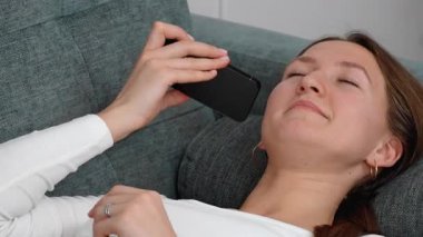 Uzun kahverengi saçlı, beyaz tişörtlü genç bir kadın gri bir kanepeye uzanıyor ve akıllı telefon kullanıyor, ciddi ile mutlu arasında yüz ifadeleri sergiliyor ve gülüyor.