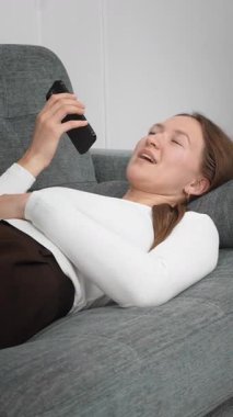 Beyaz gömlekli ve kahverengi pantolonlu genç bir kadın rahat koltukta uzanmış akıllı telefonu sağ elinde tutuyor ve gülerek sesli mesaj kaydediyor. Dikey video