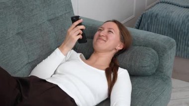 Beyaz gömlekli ve kahverengi pantolonlu genç bir kadın rahat koltukta uzanmış akıllı telefonu sağ elinde tutuyor ve sesli mesaj kaydediyor gülümsüyor ve gülüyor..