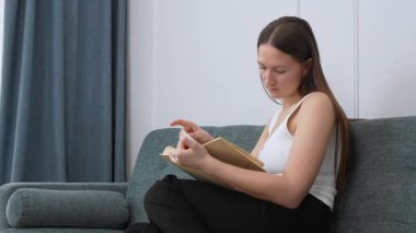 Rahat genç bir kadın, gri koltukta dinleniyor, derin bir yoğunlukla kitap okuyor, sayfaları çeviriyor, hikayenin içeriğini sakin bir ev ortamında inceliyor.