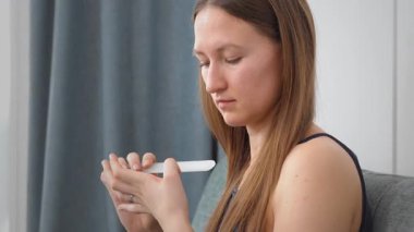 Çekici, gülümseyen genç bir kadın kanepede otururken tırnaklarını cam bir tırnak törpüsüyle parlatıyor. Yeni Güzellik Ürünleri Kullanan Güzel Kadın, Rahatlatıcı Ev, Keyifli Hafta Sonu. Günlük tırnak bakımı kavramı