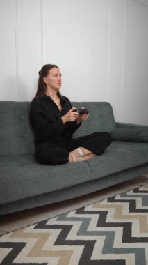 Genç bir kadın rahat bir kanepede oturuyor, kablosuz kontrolörle yoğun bir video oyunu oynuyor, rekabetçi oyun oturumunda duygularını dinamik bir şekilde ifade ediyor. Dikey video