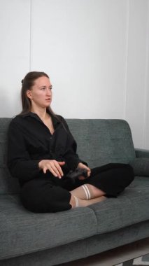 Genç bir kadın rahat bir kanepede oturuyor, kablosuz kontrolörle yoğun bir video oyunu oynuyor, rekabetçi oyun oturumunda duygularını dinamik bir şekilde ifade ediyor. Dikey video