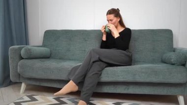 Huzurlu genç bir kadın yeşil bardaktan sıcak bir yudum yudumlarken rahat bir koltukta dinleniyor huzurlu bir ev hayatı yaşıyor.