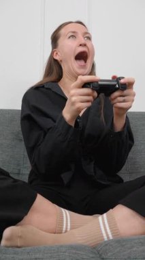 Koltuğunda oturan, elinde kumanda kolu olan mutlu genç bir kadın oturma odasındaki kanepede oturup video oyunları kazanırken heyecan verici ve çığlık çığlığa bağırıyor. dikey video