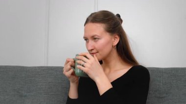 Huzurlu genç bir kadın yeşil bardaktan sıcak bir yudum yudumlarken rahat bir koltukta dinleniyor huzurlu bir ev hayatı yaşıyor.