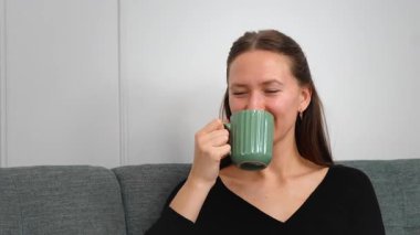 Huzurlu genç bir kadın yeşil bardaktan sıcak bir yudum yudumlarken rahat bir koltukta dinleniyor huzurlu bir ev hayatı yaşıyor..