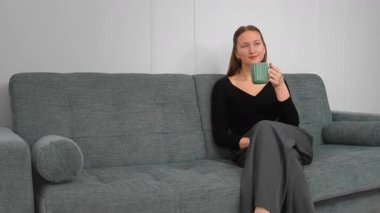 Huzurlu genç bir kadın yeşil bardaktan sıcak bir yudum yudumlarken rahat bir koltukta dinleniyor huzurlu bir ev hayatı yaşıyor..