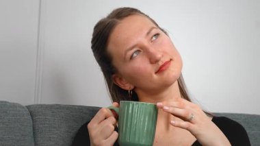 Huzurlu genç bir kadın yeşil bardaktan sıcak bir yudum yudumlarken rahat bir koltukta dinleniyor huzurlu bir ev hayatı yaşıyor..
