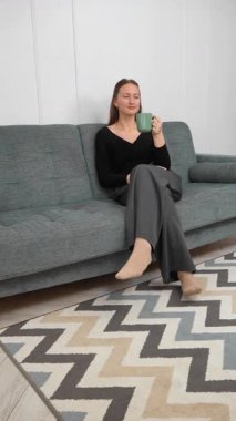 Huzurlu genç bir kadın yeşil bardaktan sıcak bir yudum yudumlarken rahat bir kanepede dinleniyor, huzurlu ev hayatı anları yakalıyor. dikey video