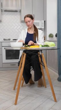 Genç ve güzel bir kadın besleyici yemekler pişiriyor zarif modern mutfağın şeffaf cam masasında renkli sebzeler kesiyor sağlıklı yaşam tarzı iş salatasına taze bir yaklaşımı somutlaştırıyor. dikey video