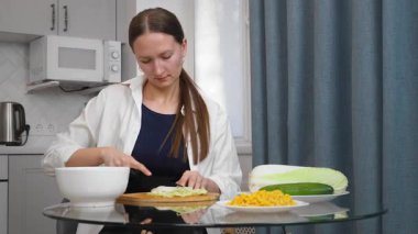 Modern mutfaktaki yuvarlak cam masada oturan sıradan giysili bir kadın evde sağlıklı yemekler hazırlarken beyaz kâse ile yemek çubuklarını karıştırıyor. Salata için sebze doğruyor.