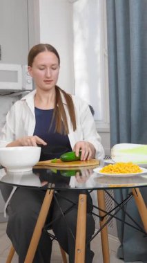 Modern mutfaktaki yuvarlak cam masada oturan sıradan giysili bir kadın evde sağlıklı yemekler hazırlarken beyaz kâse ile yemek çubuklarını karıştırıyor. Salata videosu için sebze doğruyor