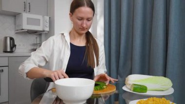 Modern mutfaktaki yuvarlak cam masada oturan sıradan giysili bir kadın evde sağlıklı yemekler hazırlarken beyaz kâse ile yemek çubuklarını karıştırıyor. Salata için sebze doğruyor.
