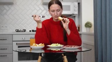Sağlıklı sosis ve salatanın tadını çıkaran genç bir kadın. Parlak, modern bir mutfakta cam masada oturmuş besleyici yemeğin tadını çıkarırken içeriğini dile getiriyor.
