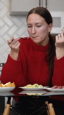 Sağlıklı sosis ve salatanın tadını çıkaran genç bir kadın. Parlak, modern bir mutfakta cam masada oturmuş besleyici yemeklerden keyif alırken dikey videolardan zevk alıyor.