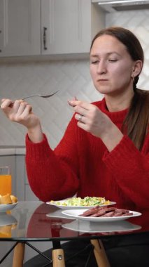 Kırmızı kazaklı genç bir kadın modern mutfağında cam bir masada otururken ekmek ve sosisli sebze salatası yiyor. Bir bardak portakal suyunun yanında. Dikey video