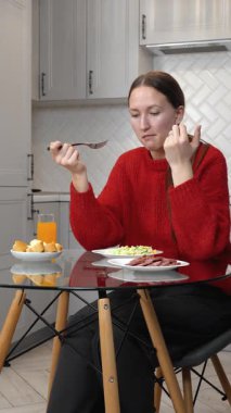 Kırmızı kazaklı genç bir kadın modern mutfağında cam bir masada otururken ekmek ve sosisli sebze salatası yiyor. Bir bardak portakal suyunun yanında. Dikey video