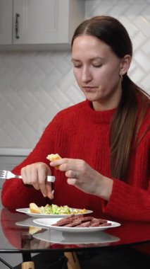 Kırmızı kazaklı genç bir kadın modern mutfağında cam bir masada otururken ekmek ve sosisli sebze salatası yiyor. Bir bardak portakal suyunun yanında. Dikey video