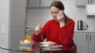 Kırmızı kazaklı genç bir kadın modern bir mutfakta cam bir masada oturmuş, dilimlenmiş sosis, salata, ekmek ve portakal suyunun tadını çıkarıyor.