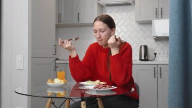 Kırmızı kazaklı genç bir kadın modern bir mutfakta cam bir masada oturmuş, dilimlenmiş sosis, salata, ekmek ve portakal suyunun tadını çıkarıyor.