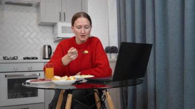 Kırmızı kazak giyen genç bir kadın, modern bir mutfakta cam bir masada oturur, laptopuyla uğraşırken aynı zamanda basit bir kahvaltı, salata, soğuk et ve portakal suyunun tadını çıkarır.