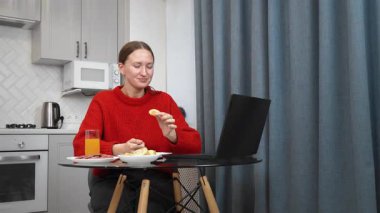 Kırmızı kazak giyen genç bir kadın mutfak masasında oturmuş, laptopunu karıştırırken kahvaltı yapıyor ve rahat ve rahat bir şekilde evde sabahın tadını çıkarıyor.