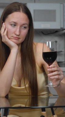 Yalnız bir kadın mutfak masasında oturmuş, uzaktan bakarken sessizce kırmızı şarap içiyor, kişisel mücadeleleri ve duygusal zorlukları yansıtıyor. Dikey video