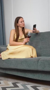 Kanepede dinlenen, kırmızı şarap içen, evde huzurlu bir gece geçirirken akıllı telefonlara bakan, sakin bir ev hayatı ve kişisel boş zamanlarında dikey video çeken bir kadın.