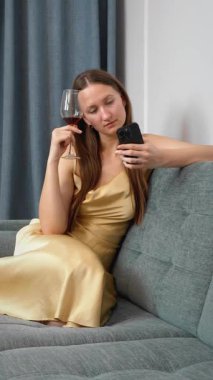 Sarı elbiseli profesyonel şarap garsonu, akıllı telefon uygulaması kullanarak mavi perdeye karşı koltukta otururken mobil teknolojiyle modern şarap uzmanlığı sergiliyor.