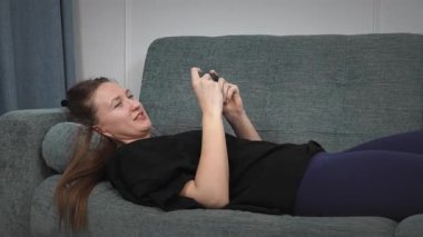 Kadın rahat bir koltukta uzanıyor, heyecanla mobil oyun oynuyor, evde boş zamanlarında neşeyle akıllı telefonu havaya fırlatarak zaferi kutluyor.