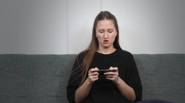 Oturma odasındaki kanepede rahat bir şekilde uzanan genç bir kadın, hareketli oyunlara çok dalmış, oynarken yüz ifadeleri dinamik bir şekilde değişiyor, saf zevk ve dijital eğlence sergiliyor.