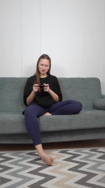 Kadın, oturma odasındaki gri bir kanepede dikey bir şekilde otururken çeşitli ifadeler ve tepkiler göstererek mobil oyunlar oynamayı seviyor.