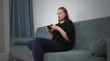 Oturma odasındaki kanepede rahat bir şekilde uzanan genç bir kadın, hareketli oyunlara çok dalmış, oynarken yüz ifadeleri dinamik bir şekilde değişiyor, saf zevk ve dijital eğlence sergiliyor.