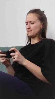 Oturma odasındaki kanepede rahat bir şekilde uzanan genç bir kadın, hareketli oyunlara çok dalmış, oynarken yüz ifadeleri dinamik bir şekilde değişiyor, saf zevk ve dijital eğlence sergiliyor.