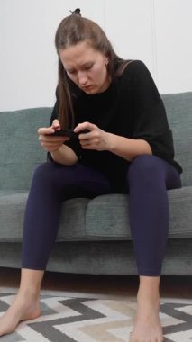 Genç bir kadın rahat bir koltukta çıplak ayakla oturuyor, akıllı telefonuyla oyun oynuyor, yüz ifadesi odaklanmaktan eğlenmeye ve dikey video seyretmeye geçiyor.