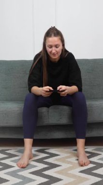 Genç bir kadın rahat bir koltukta çıplak ayakla oturuyor, akıllı telefonuyla oyun oynuyor, yüz ifadesi odaklanmaktan eğlenmeye ve dikey video seyretmeye geçiyor.