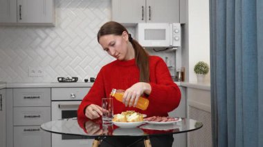 Genç bir kadın sağlıklı bir öğle yemeği yiyor. Sosis ve salata. Parlak modern bir mutfakta cam masada oturmuş, cam bardağa portakal suyu döküyor.