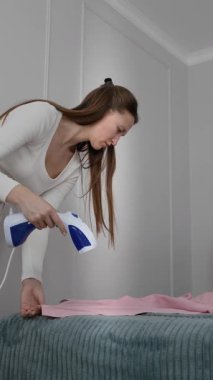 Ev hanımı rutin işlerle uğraşıyor. Ev odasındaki yataktaki kıyafetleri, el yapımı bir buharlı makineyle ütülüyor. Günlük ev işleri ve ev bakımının dikey videosunu gösteriyor.