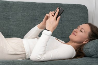 Genç kadın rahat bir şekilde kanepede uzanıyor, akıllı telefonunu huzurlu bir ifadeyle kullanıyor, modern evinde bir anlık rahatlama ve dijital bağlantının tadını çıkarıyor.