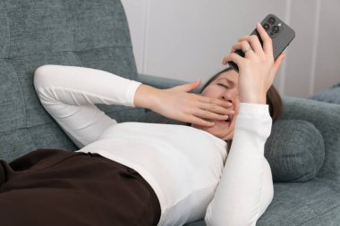 Yorgun bir kadın kanepede uzanıyor, bir eliyle ağzını kapatırken esniyor, diğer eliyle akıllı telefonunu tutuyor ve bitkin bir şekilde ekrana bakıyor.
