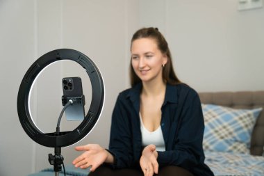 Etkilenen genç kadın, yatak odasında bir yatakta otururken el kol hareketi yaparak, halka ışığı ve akıllı telefon kullanarak sosyal medya için video içeriği kaydediyor.