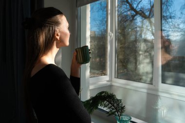 Sıcak bir içecekten zevk alan genç bir kadın güneşli bir günde pencereden dışarı bakar, şehir manzarası ya da doğa manzarası seyreder.
