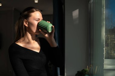 Siyah bluzlu genç bir kadın pencerenin kenarındaki yeşil bardaktan yudumlarken huzurlu bir anın tadını çıkarıyor. Güneş ışığı yavaşça yüzünü aydınlatıyor ve sakin bir atmosfer yaratıyor.