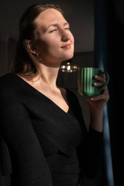 Kapalı gözleri ve sakin bir ifadeyle, genç bir kadın sabah içeceğinin sıcaklığının tadını çıkararak, yakınlardaki bir pencereden süzülen yumuşak güneş ışığının tadını çıkarıyor. Dikey fotoğraf