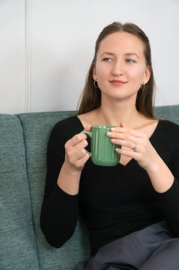 Genç bir kadın rahat bir kanepede oturmuş yeşil bir kupada sıcak bir içeceğin tadını çıkarıyor, düşünceli bir ifadeyle başka tarafa bakıyor, rahat ve rahatlatıcı bir atmosfer yaratıyor. Dikey fotoğraf
