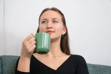 Genç bir kadın rahat bir kanepede oturmuş sıcak bir içeceğin tadını çıkarıyor, derin derin bakıyor, rahat ve düşünceli bir atmosfer yaratıyor.